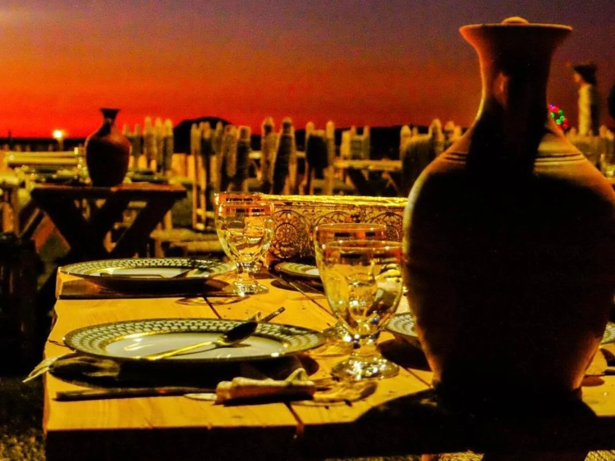 A cozy Agafay desert dinner setup in Agafay Desert with traditional Moroccan dinnerware, glassware, and a stunning sunset backdrop.