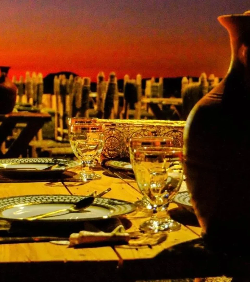 A cozy Agafay desert dinner setup in Agafay Desert with traditional Moroccan dinnerware, glassware, and a stunning sunset backdrop.