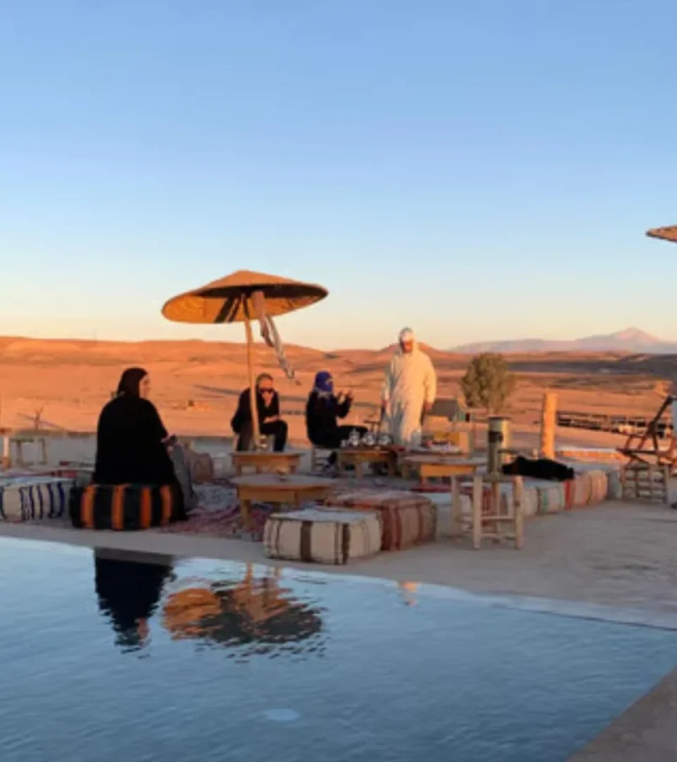 Guests relaxing at a Sahara camp in Morocco, sitting by the pool with desert views, traditional decor, and a peaceful ambiance.