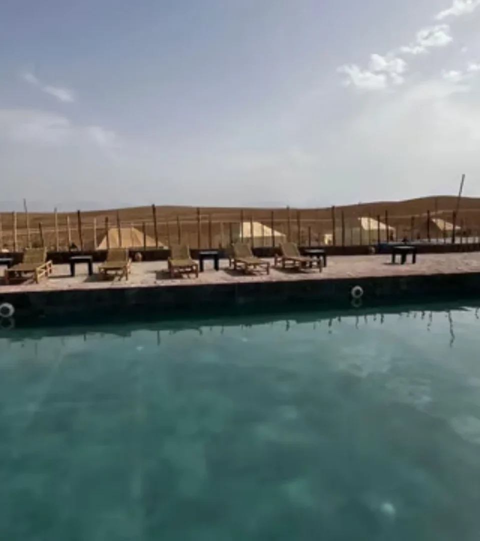 A luxurious Morocco Sahara Desert camp with a peaceful pool, sun loungers, and scenic desert tents in the background.