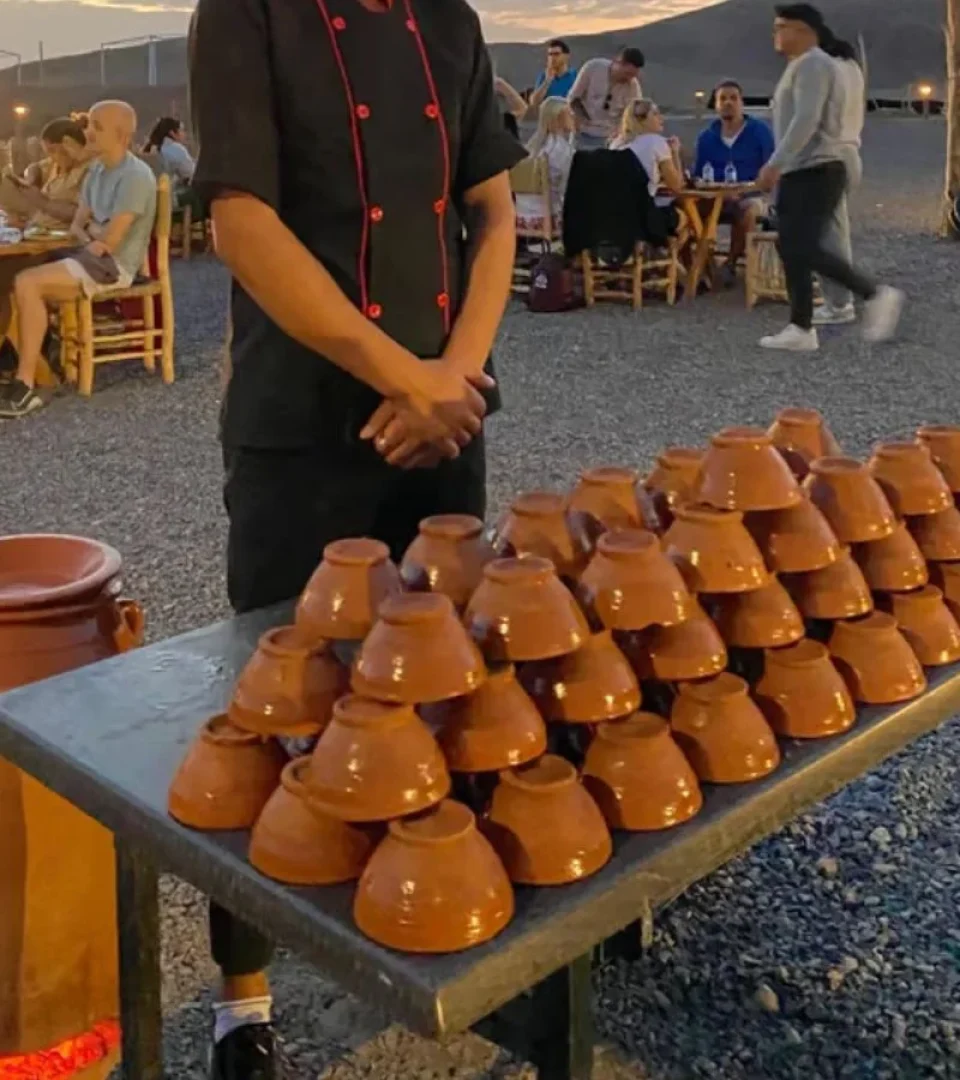 A chef stands beside a stacked display of traditional Moroccan tagine bowls at a Marrakech Agafay Desert dinner in Berberkamp and camel ride event.