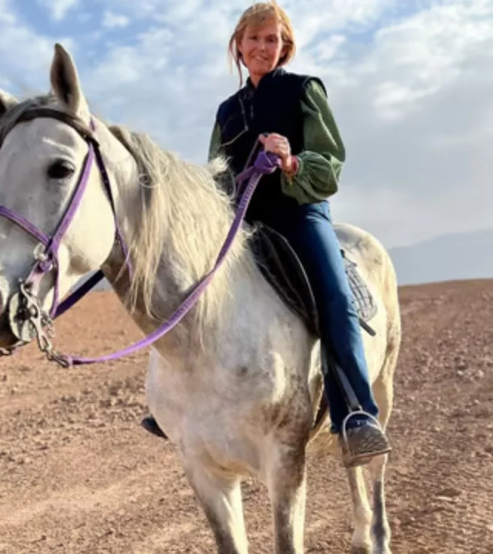 A smiling rider enjoying horse riding in Marrakech, sitting on a majestic white horse with desert mountains in the background.