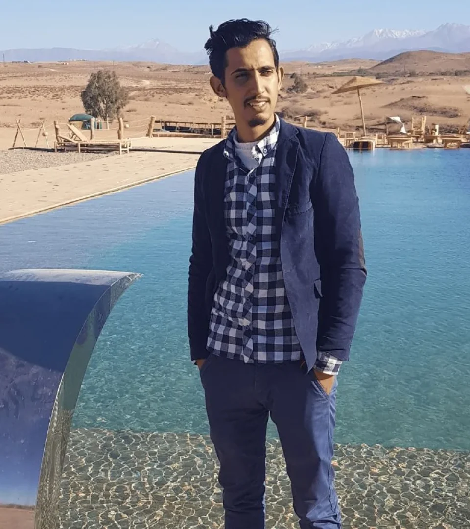 A man standing by the pool at a Sahara Desert luxury camp, dressed stylishly with scenic desert views in the background.